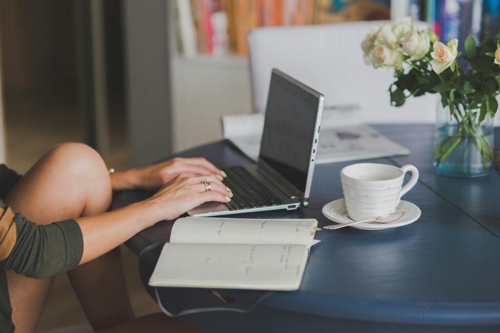 A person typing on a laptop at a dark blue table with an open notebook, a white coffee cup on a saucer, and a vase of white roses. The setting is cozy and well-lit, with bookshelves and a magazine in the background.