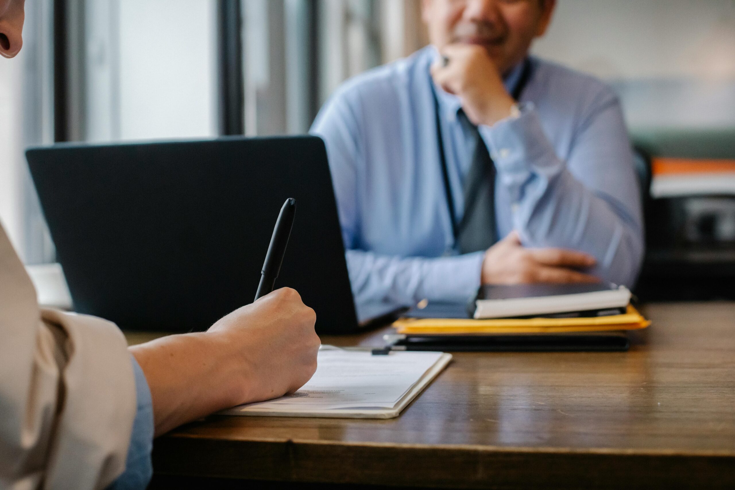 A close-up of a person writing with a black pen on a notepad during a professional meeting, with a smiling man in a blue shirt sitting across the table. A laptop, notebooks, and documents are placed on the wooden desk.