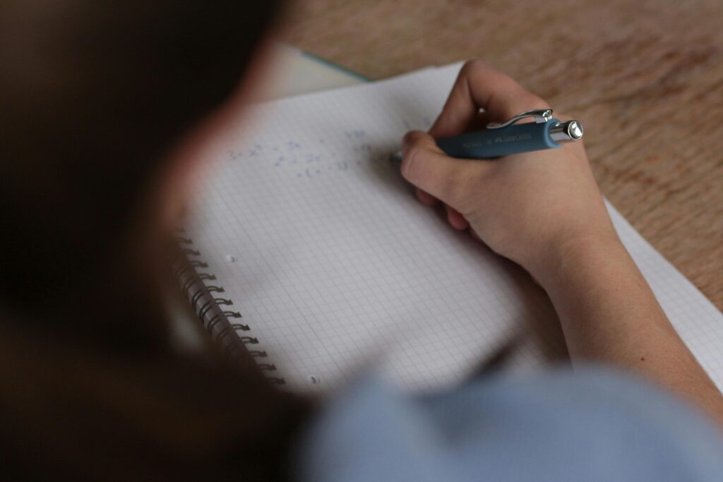 pexels-louis-bauer-79024-249360 The image shows a person writing mathematical equations in a spiral notebook with a grid (graph paper) layout. The person is using a blue mechanical pencil or pen from the brand Faber-Castell. The equations appear to involve algebraic expressions, possibly solving for a variable like 𝑥 x. The setting looks like a study or homework environment, with a wooden surface underneath the notebook. The person's head and shoulder are slightly blurred in the foreground, focusing attention on the writing hand.